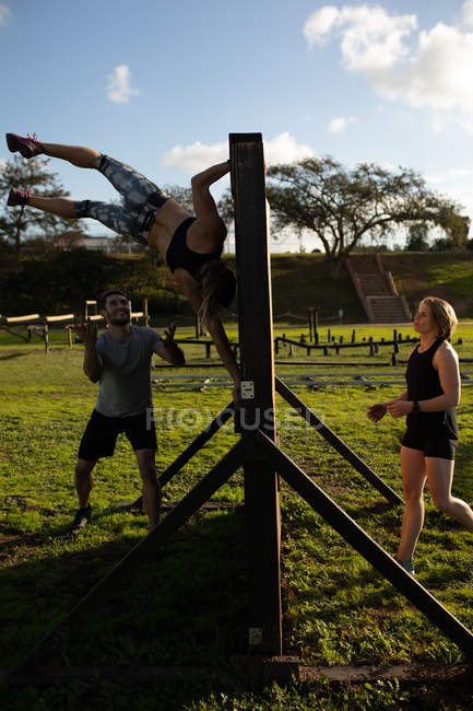 Rear view of a young Caucasian woman climbing down from a wall, a young Caucasian man with his arms held out to catch her, and a young Caucasian woman waiting to climb the wall at an outdoor gym during a bootcamp training session — Stock Photo