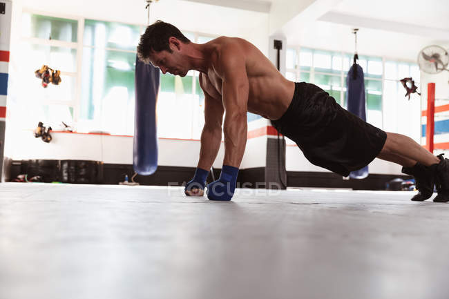 Vista lateral de un joven boxeador caucásico haciendo flexiones en un gimnasio de boxeo - foto de stock