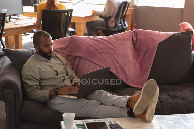 Vista frontal de cerca de un joven afroamericano sentado en un sofá con los pies en una mesa usando una computadora portátil en la sala de estar de una oficina creativa, con un colega trabajando de fondo - foto de stock