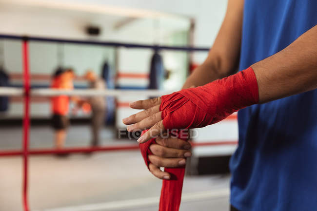 Vista lateral sección media de boxeador masculino en un anillo de boxeo envolviendo sus manos mientras otro joven está boxeando en un fondo - foto de stock