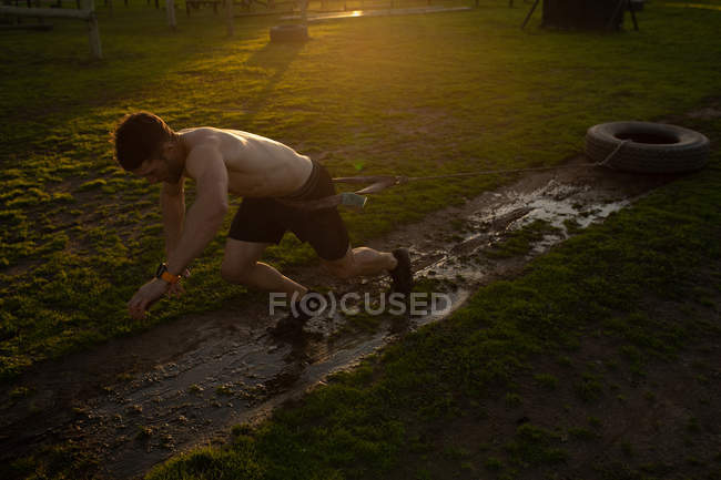 Seitenansicht eines jungen kaukasischen Mannes, der während eines Bootcamp-Trainings in einem Outdoor-Fitnessstudio einen Reifen an einem Seil um die Hüfte durch Schlamm zieht — Stockfoto