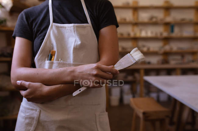 Vue de face section médiane un potier féminin portant un tablier tenant une brosse à vitrage dans un atelier de poterie — Photo de stock