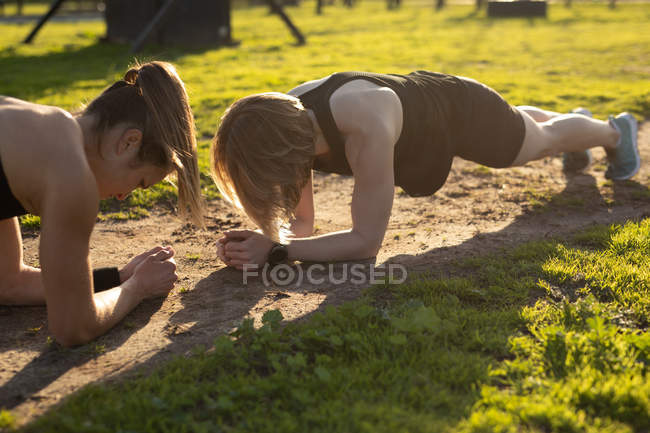 Vista lateral de duas jovens mulheres caucasianas fazendo o exercício de prancha em um ginásio ao ar livre durante uma sessão de treinamento de bootcamp — Fotografia de Stock