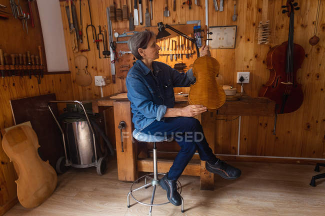 Seitenansicht einer älteren kaukasischen Geigenbauerin mit einer Geige in ihrer Werkstatt, im Hintergrund hängen Werkzeuge an der Wand — Stockfoto