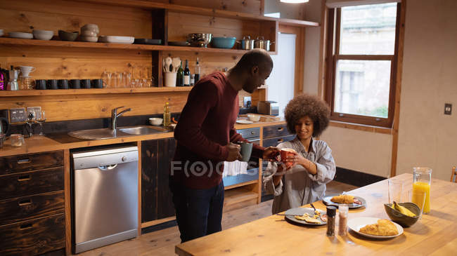 Вид збоку молодого афро-американського людини стоячи і говорити, як він проходить чашку кави до свого партнера, посміхаючись молодих змішаних гонка жінка, сидячи за своїм кухонним столом і їдять вдома — стокове фото