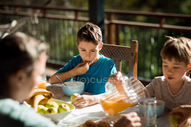 Vista frontal de cerca de dos niños caucásicos pre adolescentes sentados en una mesa disfrutando de un desayuno familiar en un jardín - foto de stock