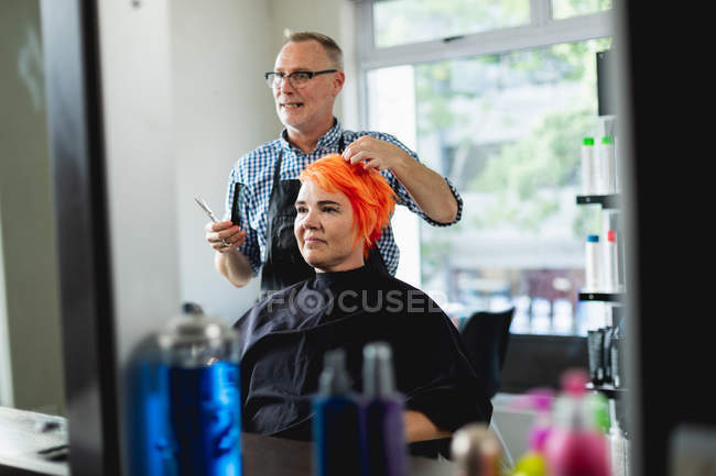 Front view of a middle aged Caucasian male hairdresser and a young Caucasian woman having her hair colored bright red and cut in a hair salon, reflected in a mirror — Stock Photo