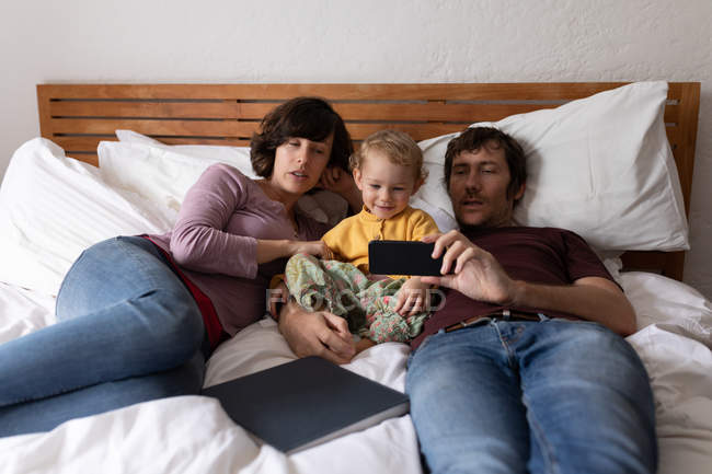 Front view of a young Caucasian father and mother lying on a bed and using a smartphone with their baby — Stock Photo