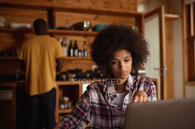 Vista frontal de cerca de una joven mujer de raza mixta usando un ordenador portátil sentado en la mesa de su cocina, su pareja, un joven afroamericano, está de pie con la espalda a la cámara en el fondo - foto de stock