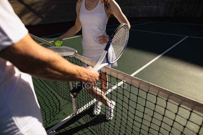 Vista frontal close-up de mulher e um homem segurando raquetes e uma bola em um campo de tênis em um dia ensolarado — Fotografia de Stock