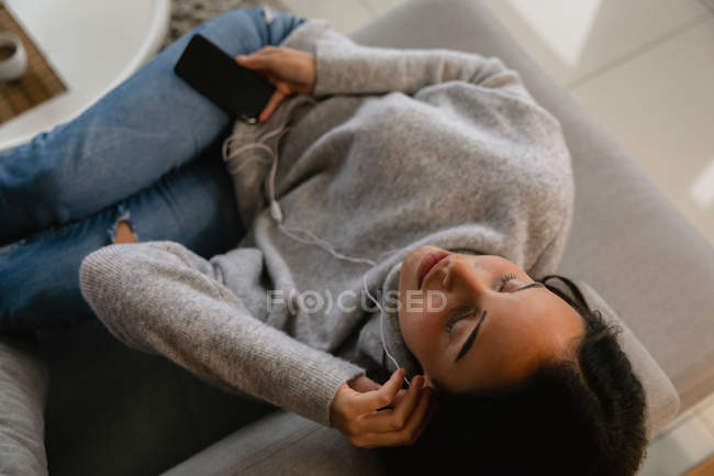 Підвищений вид молодий Кавказький жінка брюнетка лежачи на дивані з закритими очима прослуховування музики за допомогою смартфона і навушники — стокове фото