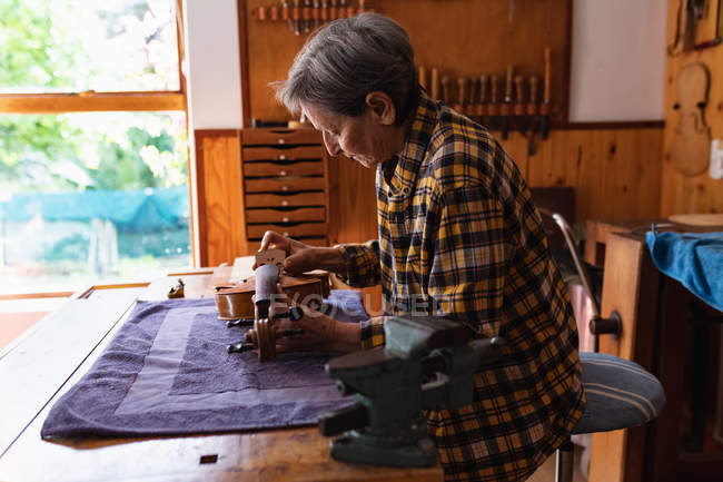 Вид збоку старших кавказьких жінок luthier працює над тілом скрипки в її майстерні з інструментами звисають на стіні у фоновому режимі — стокове фото