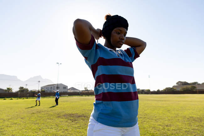 Front view of a young adult mixed race female rugby player standing on a rugby pitch fastening her headguard, with her teammates in the background — Stock Photo