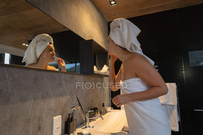 Seitenansicht einer jungen kaukasischen Frau mit in ein Handtuch gewickelten Haaren, die sich die Zähne putzt und in einem modernen Badezimmer in den Spiegel schaut — Stockfoto