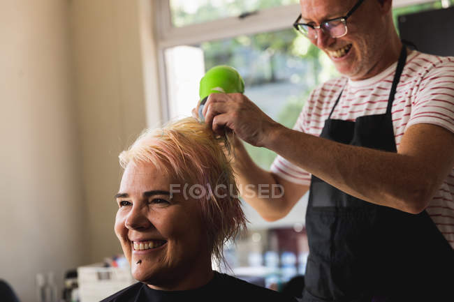 Vista lateral de perto de um cabeleireiro masculino caucasiano de meia idade e uma jovem mulher caucasiana com o cabelo seco em um salão de cabeleireiro — Fotografia de Stock