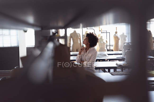 Vista lateral de una joven estudiante de moda de raza mixta mirando diseños en una pared en un estudio de la universidad de moda, con un primer plano desenfocado - foto de stock
