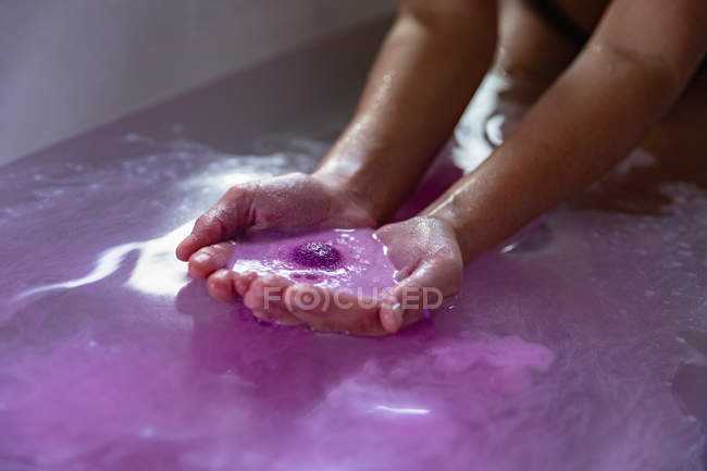 Acercamiento de las manos ventosas de una mujer en un baño con sales de baño rosadas efervescentes en el agua del baño - foto de stock