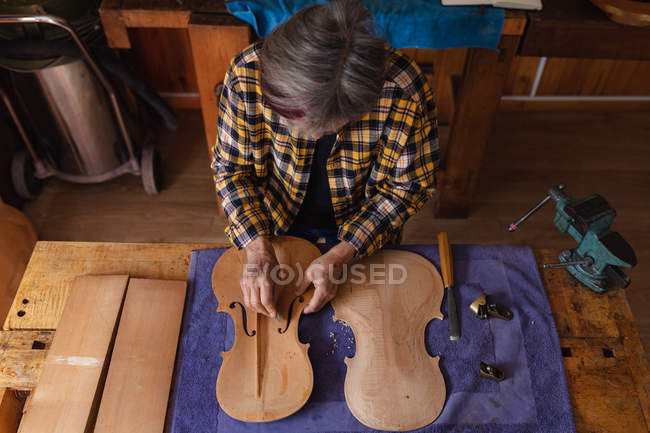 Vista de alto ángulo de una luthier femenina caucásica mayor trabajando en el cuerpo de un violín en su taller - foto de stock