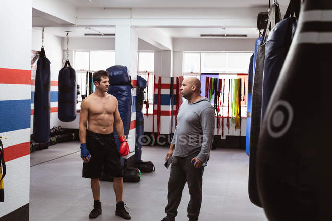 Vista frontal de un joven boxeador caucásico en un gimnasio de boxeo hablando con un entrenador masculino caucásico de mediana edad - foto de stock