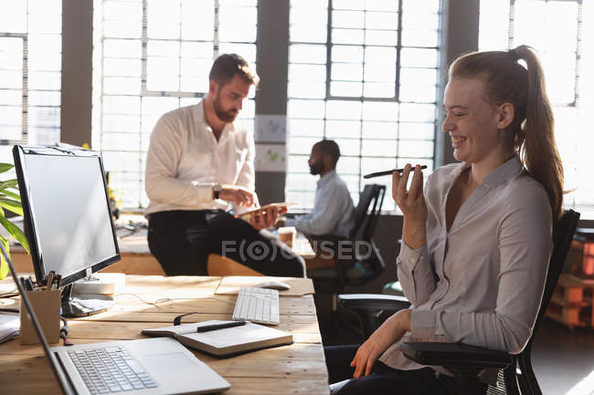 Vista lateral de cerca de una joven mujer caucásica sentada en un escritorio en una computadora hablando en un teléfono inteligente en una oficina creativa, con un joven colega caucásico sentado en un escritorio usando una tableta - foto de stock