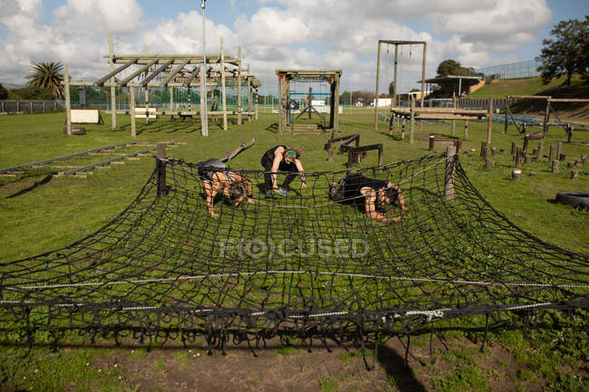 Vista frontal de dos jóvenes mujeres caucásicas y un joven hombre caucásico arrastrándose bajo una red en un gimnasio al aire libre durante una sesión de entrenamiento de campo de entrenamiento - foto de stock