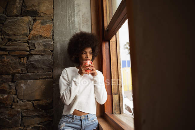 Front view close up of a young mixed race woman standing by a window, looking out and holding a cup of coffee — Stock Photo