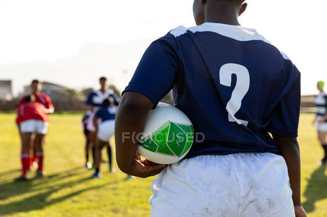 Vista trasera sección central de una joven jugadora de rugby afroamericana adulta de pie en un campo de rugby con una pelota de rugby bajo su brazo, con jugadores de ambos equipos en el fondo durante un partido de rugby - foto de stock