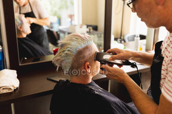 Vista laterale da vicino di un parrucchiere caucasico di mezza età e di una giovane donna caucasica che si fa tagliare i capelli in un parrucchiere, riflesso in uno specchio — Foto stock