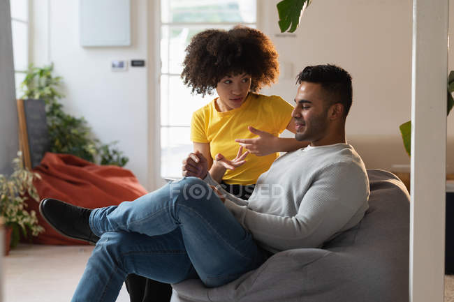 Вид спереду крупним планом молодий чоловік змішаної раси і молода змішана жінка, що сидить на мішку з квасолею, використовуючи планшетний комп'ютер і обговорюючи в творчому офісі — стокове фото