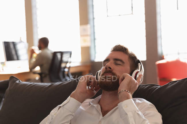 Vista frontal de cerca de un joven caucásico apoyado en un sofá con los ojos cerrados usando auriculares en la sala de estar de un negocio creativo - foto de stock
