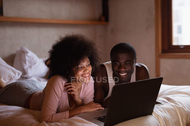 Вид спереду крупним планом молодої жінки змішана гонка і її партнер, молодий афроамериканець людина, лежачи на ліжку спираючись на лікті посміхається і дивитися ноутбук на дому — стокове фото