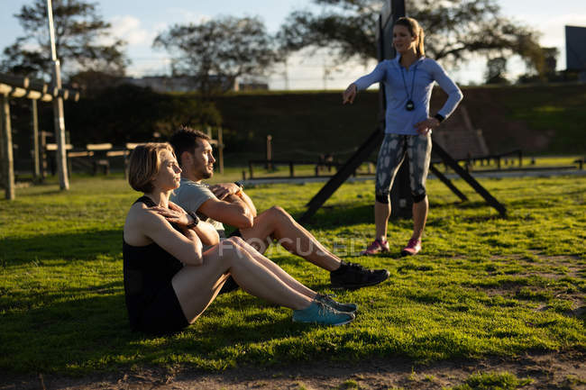 Vista lateral de una joven mujer caucásica y un joven hombre caucásico sentado en la hierba haciendo ejercicio mientras una joven instructora caucásica está hablando con ellos en un gimnasio al aire libre durante una sesión de entrenamiento de campo de entrenamiento - foto de stock