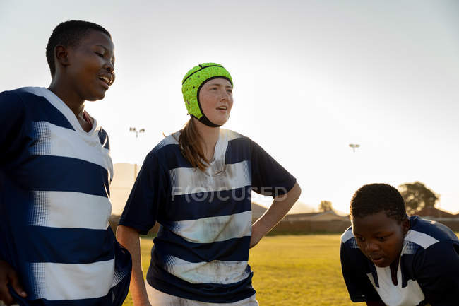 Vista frontal de dos jugadoras de rugby afroamericanas adultas jóvenes y una jugadora de rugby caucásica adulta joven con un protector de cabeza relajándose después de un partido de rugby - foto de stock