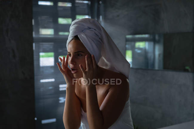 Vista frontale da vicino di una giovane donna bruna caucasica che indossa un asciugamano da bagno e con i capelli avvolti in un asciugamano, si guarda allo specchio e si massaggia il viso con le dita in un bagno moderno — Foto stock