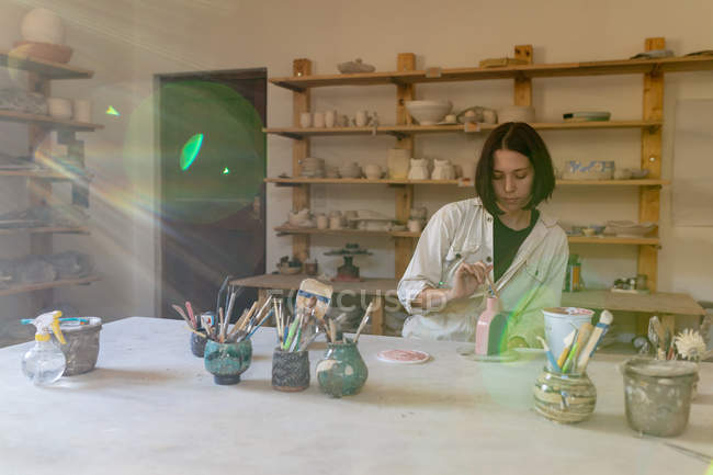 Vista frontale di una giovane ceramista caucasica seduta su un tavolo da lavoro che dipinge uno smalto colorato su una fiaschetta di argilla in uno studio di ceramica — Foto stock