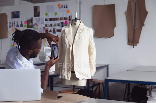 Vista lateral de un joven afroamericano estudiante de moda usando un teléfono inteligente para tomar una foto de un diseño de chaqueta en un maniquí en un estudio en la universidad de moda - foto de stock