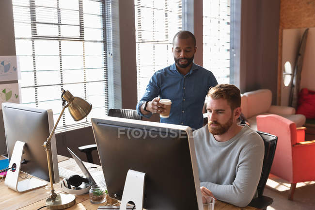 Vorderseite eines jungen afrikanisch-amerikanischen Mannes, der einen Kaffee hält und mit einem jungen kaukasischen männlichen Kollegen spricht, der an einem Schreibtisch sitzt und einen Computer in einem kreativen Büro benutzt — Stockfoto