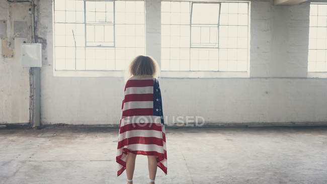 Заднього виду молода кавказька жінка з Кучеряве волосся і американський прапор над плечима, стоячи всередині порожній склад — стокове фото