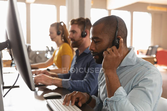Vista lateral de cerca de un joven afroamericano y una joven mujer y hombre caucásicos sentados en escritorios usando computadoras y hablando con auriculares telefónicos en una oficina creativa - foto de stock