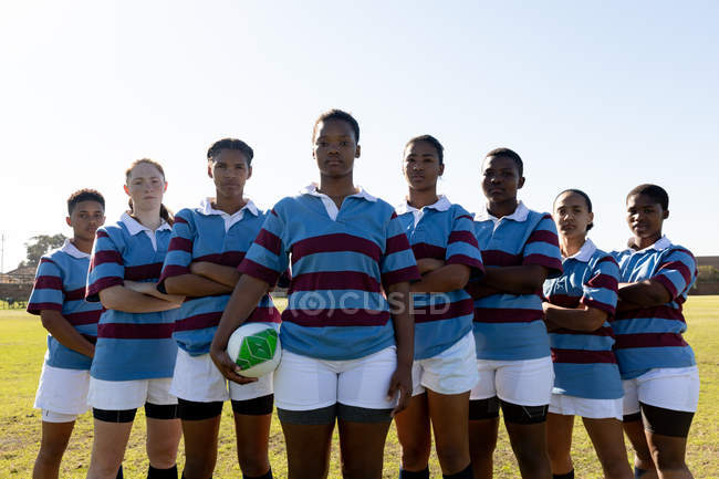 Portrait d'une équipe de jeunes joueuses de rugby multiethniques adultes debout en formation sur un terrain de rugby avec les bras croisés, regardant la caméra sur un terrain de rugby, la femme à l'avant tient une balle de rugby — Photo de stock