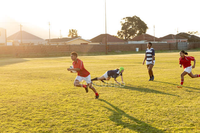 Seitenansicht einer Gruppe junger erwachsener multiethnischer Rugbyspielerinnen, die während eines Spiels auf das Spielfeld laufen und dem Spieler in Ballbesitz hinterherjagen — Stockfoto