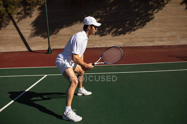 Vista lateral de un joven caucásico jugando al tenis en un día soleado, sosteniendo una raqueta y esperando la pelota - foto de stock