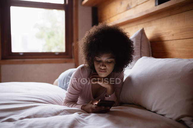 Вид спереду крупним планом молода змішана жінка, що лежить на її передній частині на ліжку, спираючись на лікті, використовуючи смартфон вдома — стокове фото