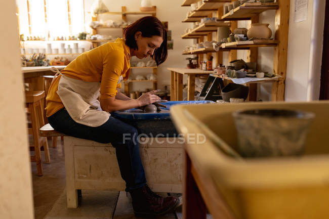 Vista lateral de uma jovem cerâmica caucasiana sentada e trabalhando com argila em uma roda de oleiros em um estúdio de cerâmica, com equipamento em primeiro plano — Fotografia de Stock