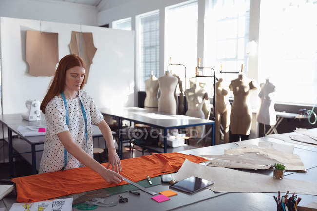 Side view of a young Caucasian female fashion student working on a design measuring orange fabric in a studio at fashion college, with mannequins in the background — Stock Photo