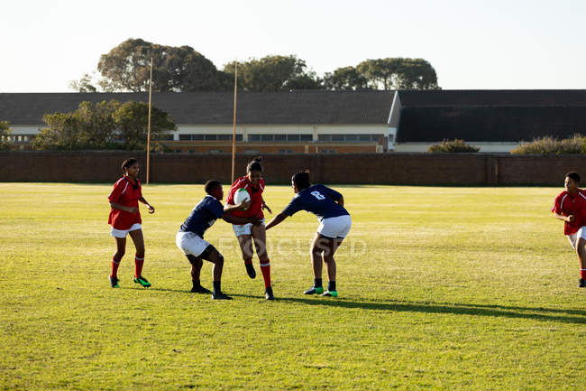 Передній вигляд групи молодих дорослих багатоетнічних гравців регбі під час матчу, з двома гравцями, які намагаються зупинити одного гравця, який біжить з м'ячем — стокове фото