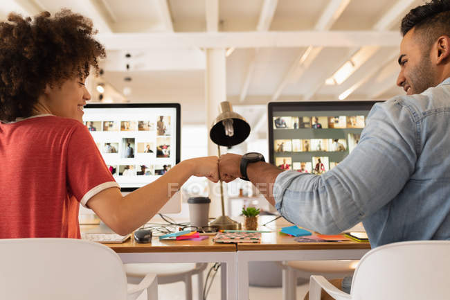Rear view close up of a young mixed race man and a young mixed race woman sitting at a desk using computers and celebrating with a fist bump at a creative office — Stock Photo