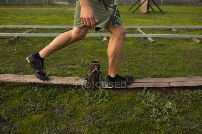 Vista lateral sección baja del hombre caminando a lo largo de postes de madera en un gimnasio al aire libre durante una sesión de entrenamiento de bootcamp - foto de stock