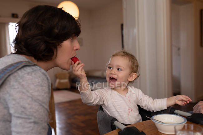 Vista lateral de cerca de una joven madre caucásica siendo alimentada con una fresa por su bebé - foto de stock
