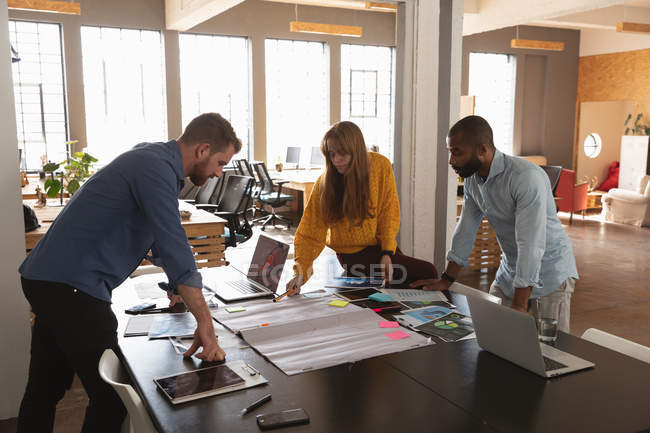 Side view of a young African American man and a young Caucasian man and woman standing and leaning over a desk working together on a project in a creative office — Stock Photo